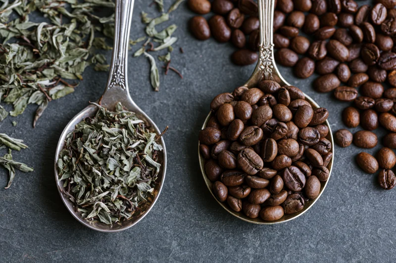 Two vintage silver spoons holding coffee beans and dried tea leaves on dark slate surface