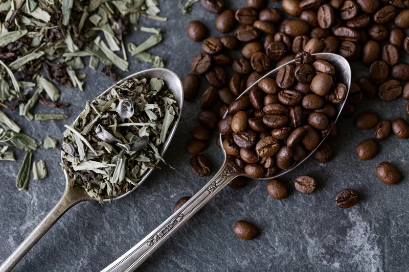 Two vintage silver spoons holding coffee beans and dried herbs on dark slate surface