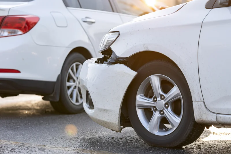 Two white cars in rear-end collision with damaged front bumper and crumpled bodywork on asphalt road