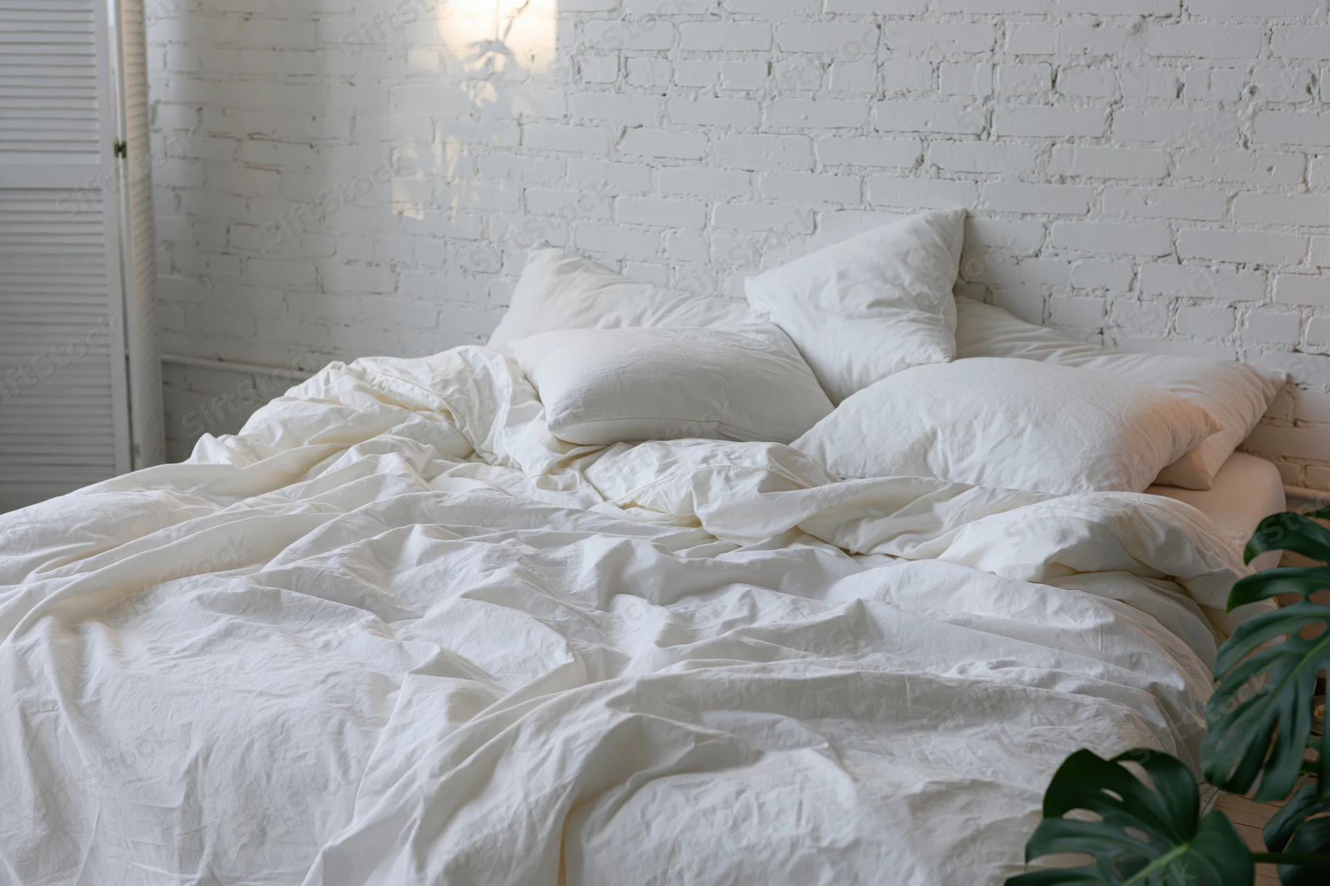 Unmade bed with rumpled white cotton sheets and four pillows against a white brick wall in a bedroom