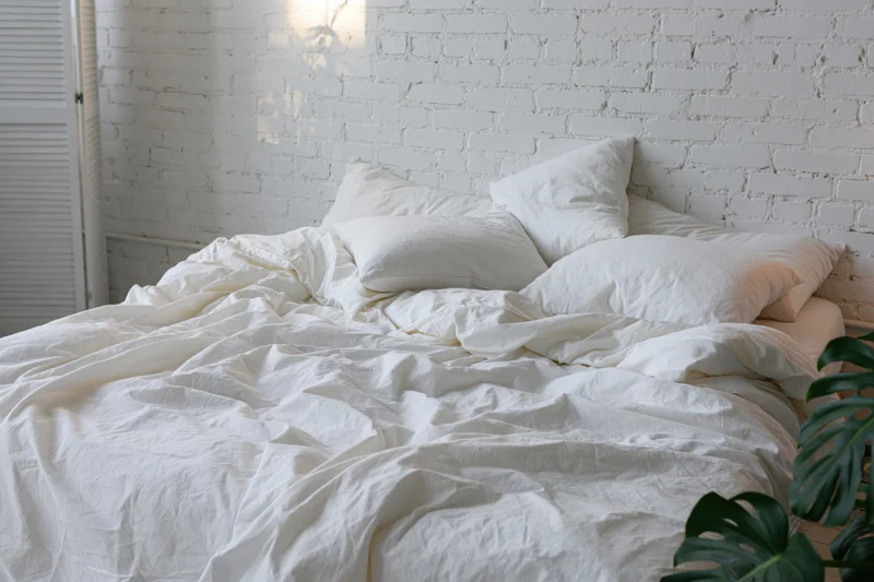 Unmade bed with rumpled white cotton sheets and four pillows against a white brick wall in a bedroom
