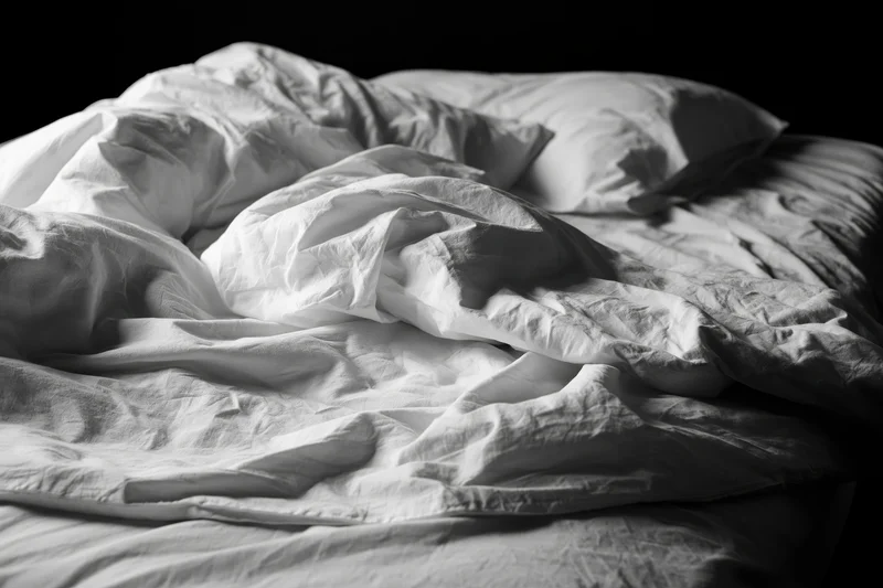 Unmade bed with rumpled white sheets and tangled pillows in a dark bedroom