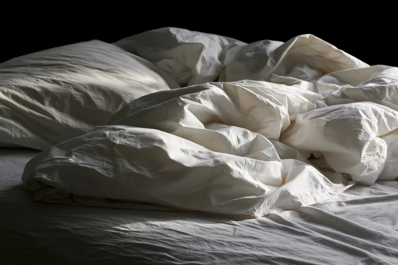 Unmade bed with rumpled white sheets and tangled pillows in a dark bedroom