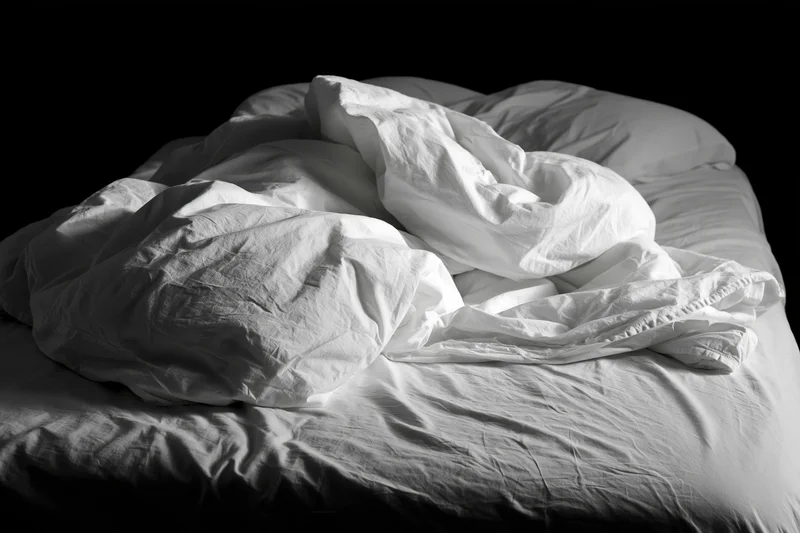 Unmade bed with rumpled white sheets and tangled pillows in a dark room