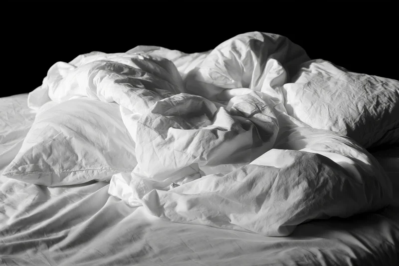 Unmade bed with rumpled white sheets and tangled pillows against black background