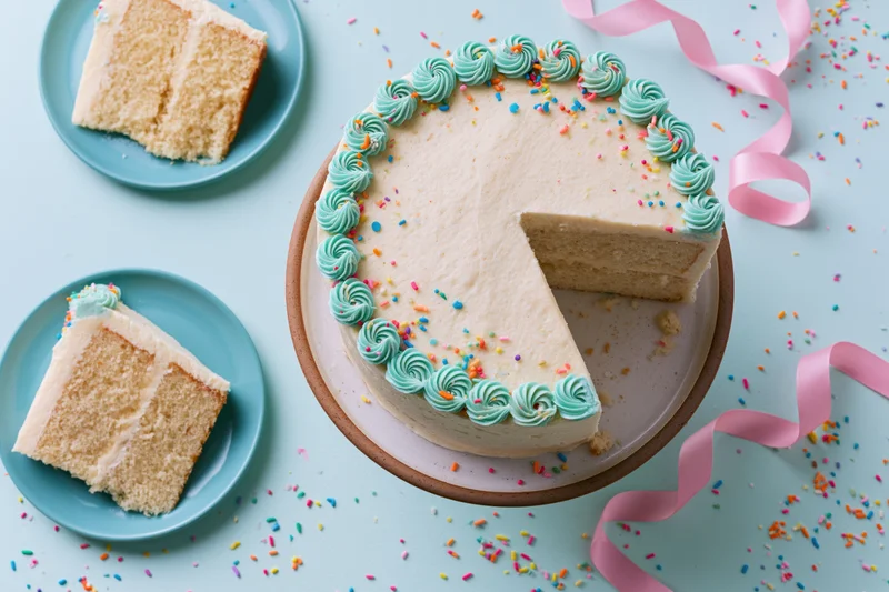 White-frosted layered birthday cake with a slice removed on a cake stand surrounded by sprinkles and pink ribbon