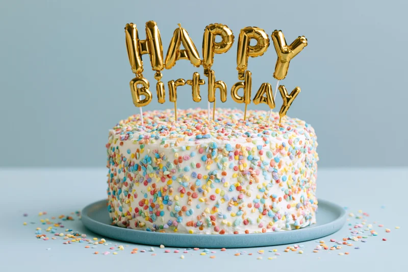 White birthday cake covered in colorful sprinkles with gold balloon letter topper on blue plate
