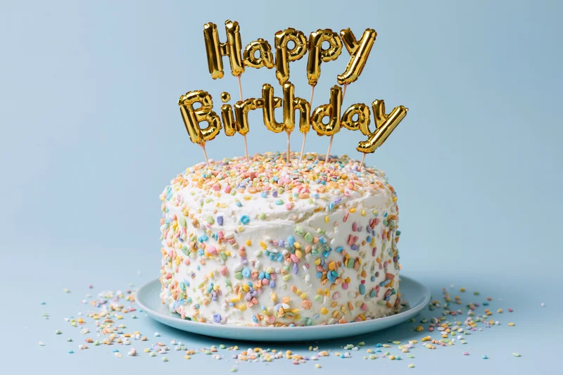 White birthday cake covered in colorful sprinkles with gold balloon lettering topper on blue background