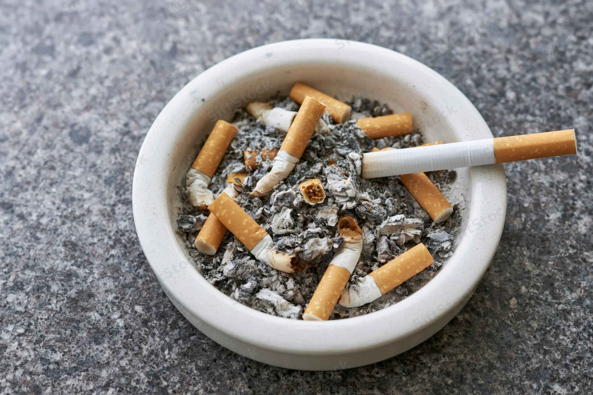White ceramic ashtray filled with cigarette butts and gray ash on a stone surface