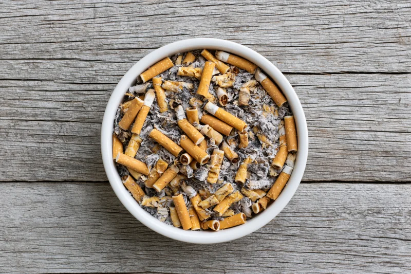 White ceramic ashtray filled with used cigarette butts and ash on wooden surface