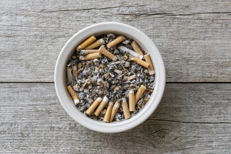 White ceramic ashtray filled with cigarette butts and ash on a weathered wooden surface