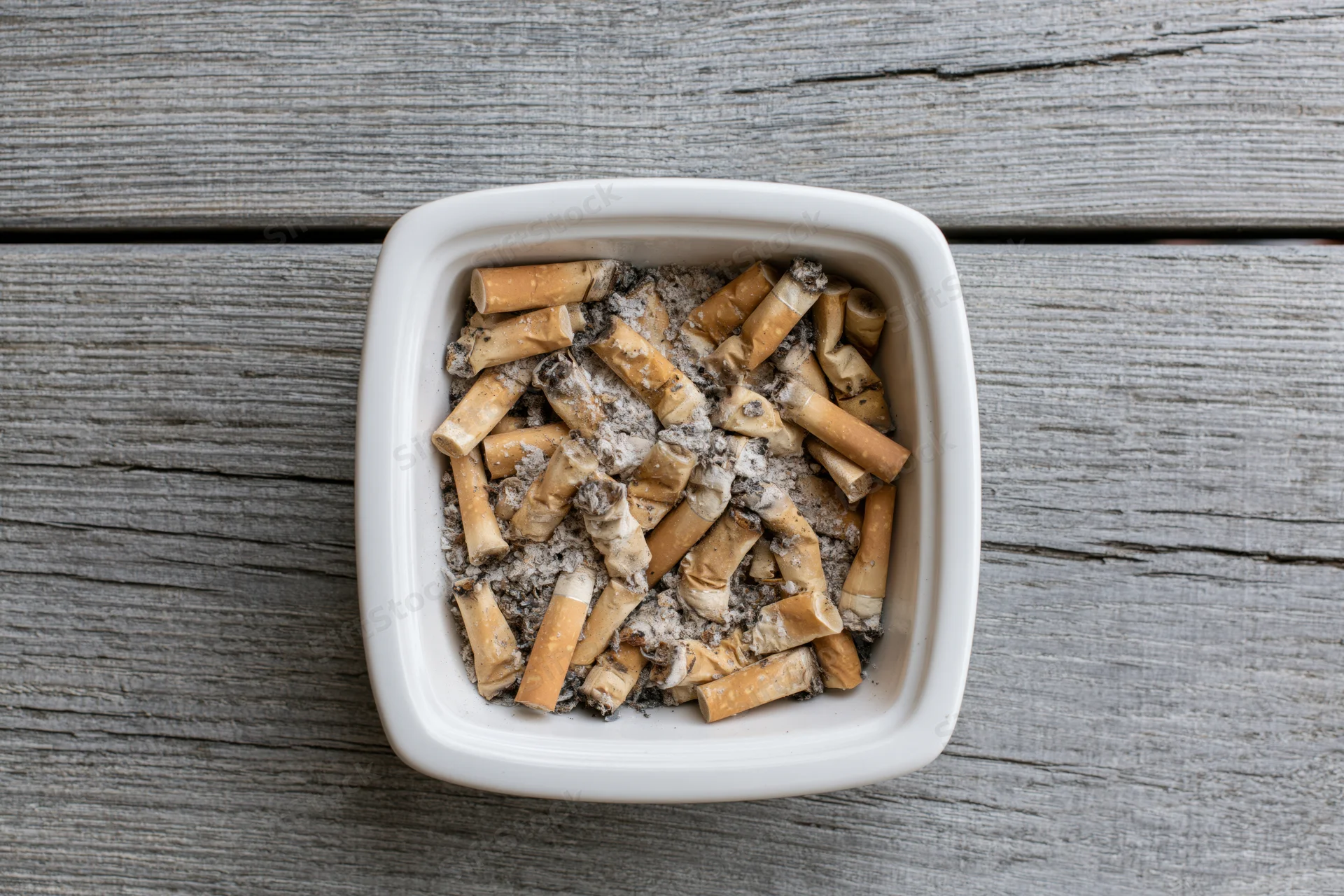 White ceramic ashtray filled with cigarette butts and ash on a gray wooden surface
