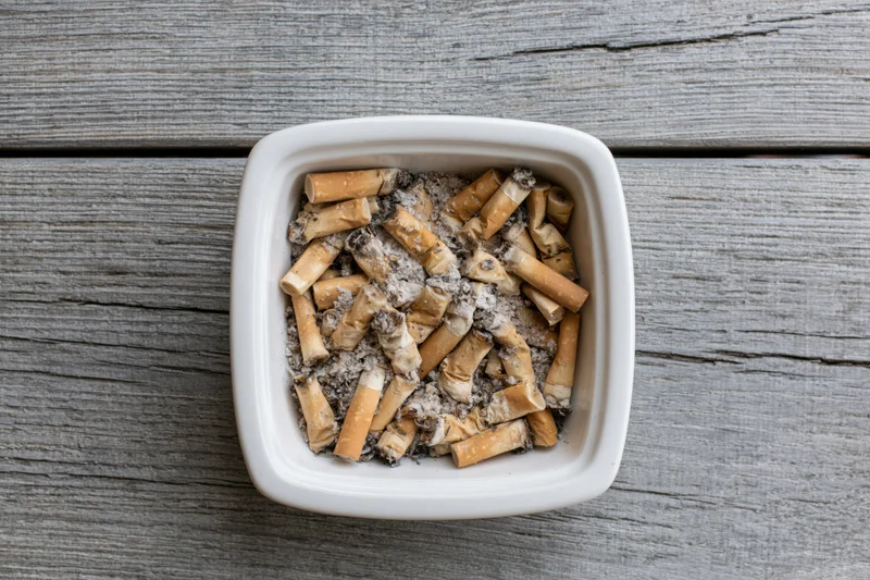 White ceramic ashtray filled with cigarette butts and ash on a gray wooden surface
