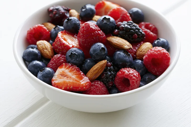 A white ceramic bowl filled with mixed berries and almonds on a light wooden surface