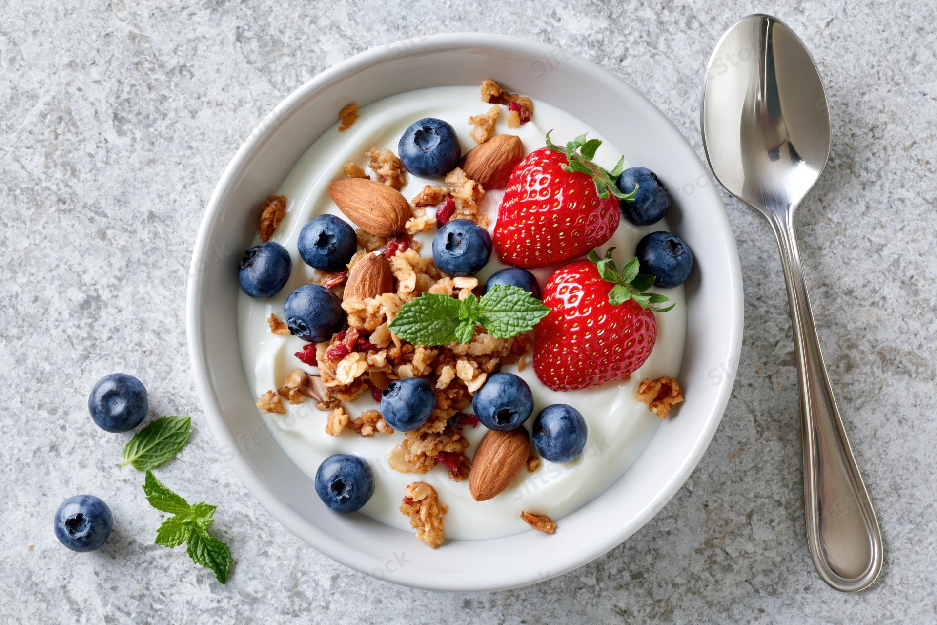 White ceramic bowl of yogurt topped with granola, blueberries, strawberries, almonds and mint, spoon beside on textured surface