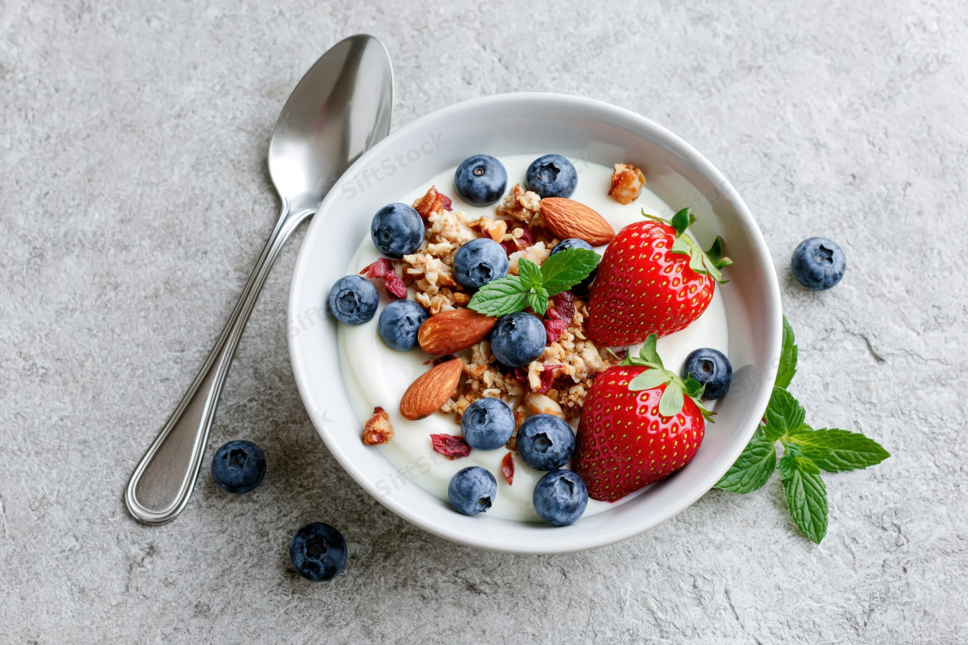 White ceramic bowl of yogurt topped with granola, blueberries, strawberries, almonds, and mint on gray surface with spoon beside it