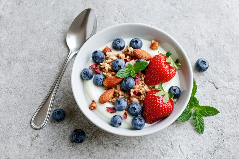 White ceramic bowl of yogurt topped with granola, blueberries, strawberries, almonds, and mint on gray surface with spoon beside it