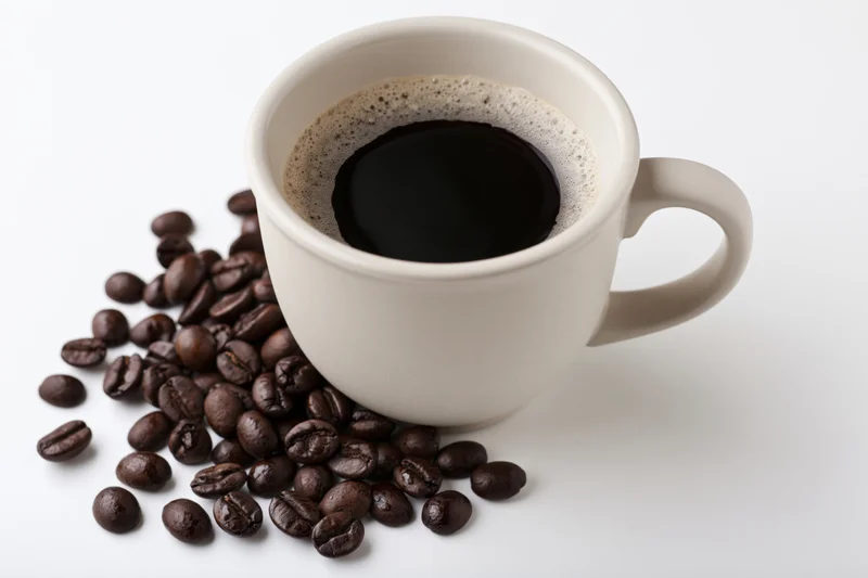 White ceramic coffee cup filled with black coffee and foam beside scattered coffee beans on white surface