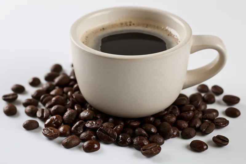 White ceramic coffee cup filled with black coffee and foam resting on scattered roasted coffee beans on a white surface