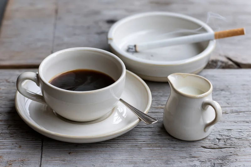 White ceramic coffee cup with espresso and spoon on saucer beside ashtray with cigarette and milk pitcher on wooden table