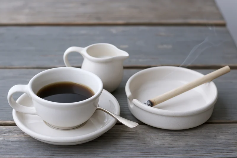 White ceramic coffee cup with espresso and spoon on saucer next to ashtray with smoking cigarette on wooden table