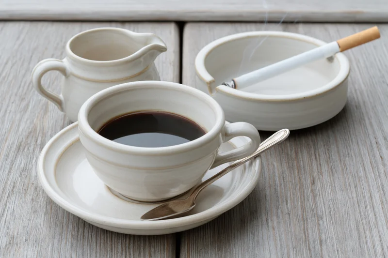 White ceramic coffee cup with espresso and spoon on saucer beside ashtray with smoking cigarette on wooden table