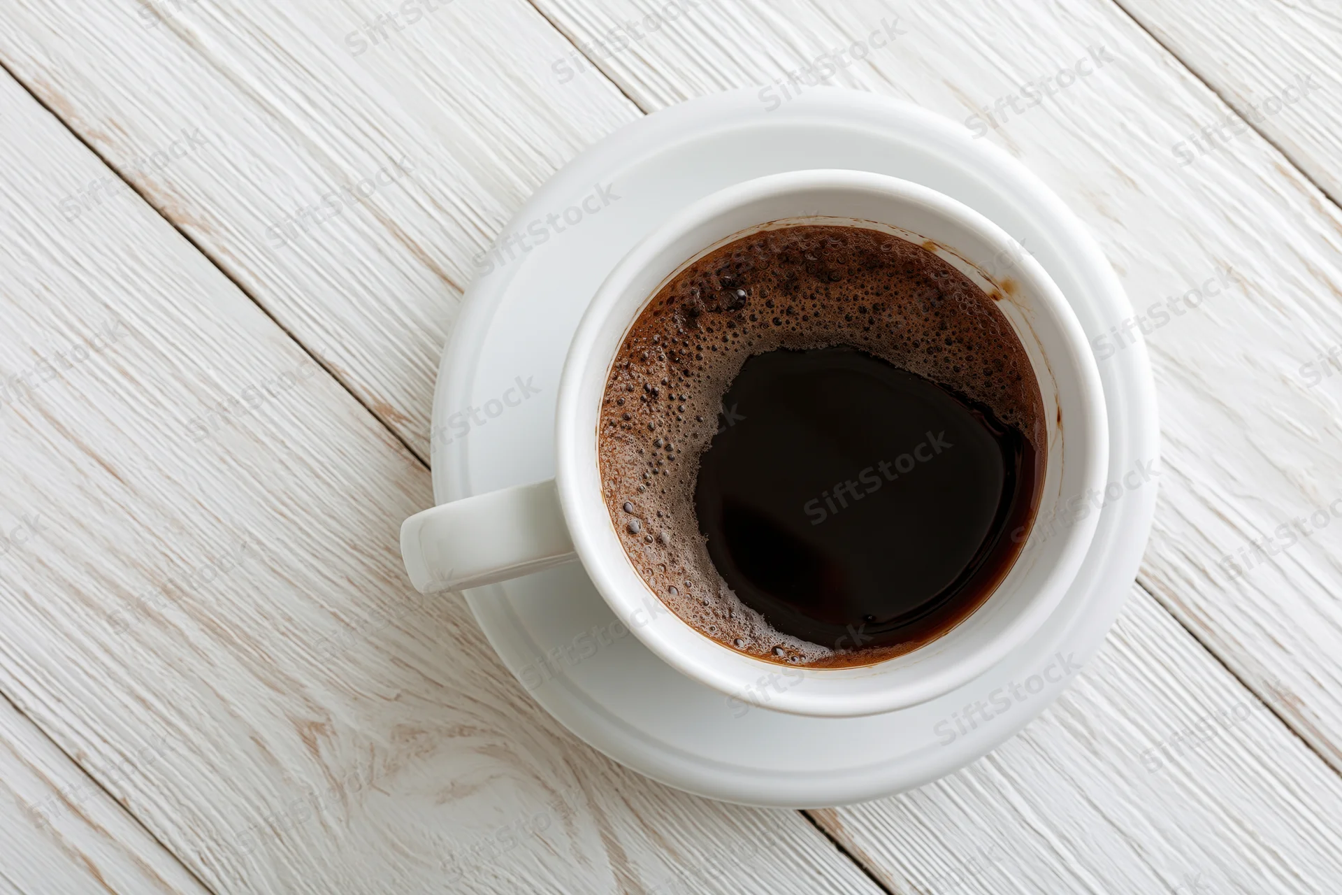 White ceramic coffee cup filled with black coffee on a saucer atop a whitewashed wooden table, overhead view