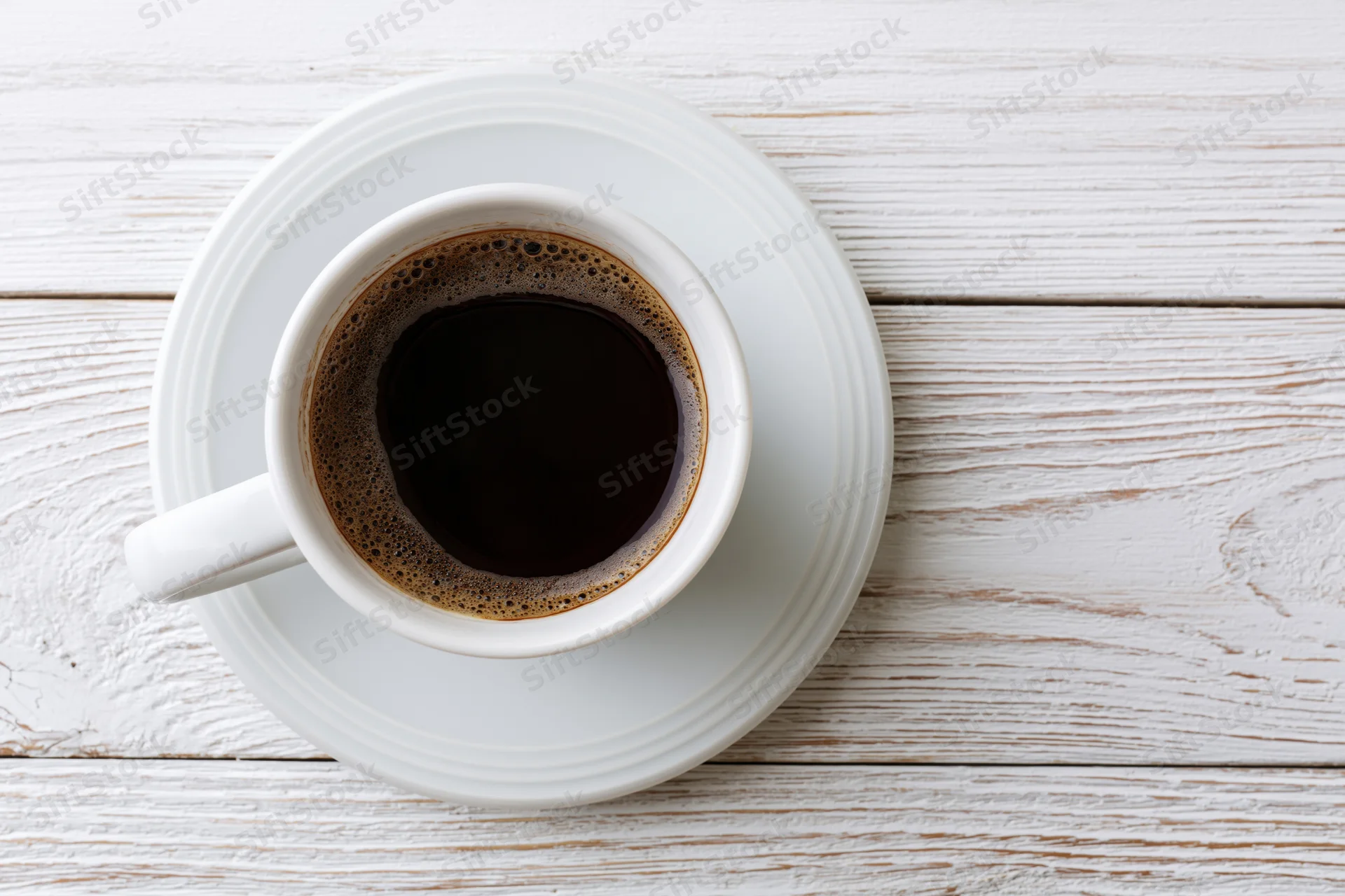 White ceramic coffee cup filled with dark coffee on a matching saucer atop a whitewashed wooden table, top-down view