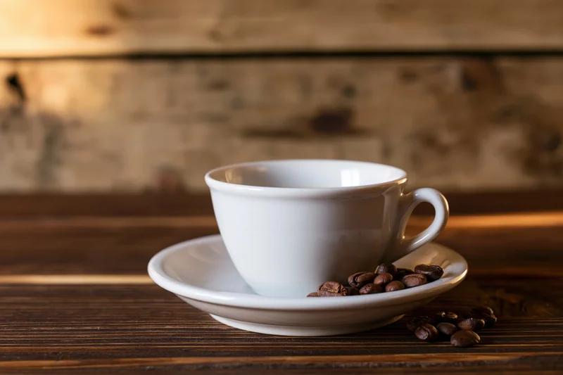 White ceramic coffee cup on saucer with roasted beans on wooden table