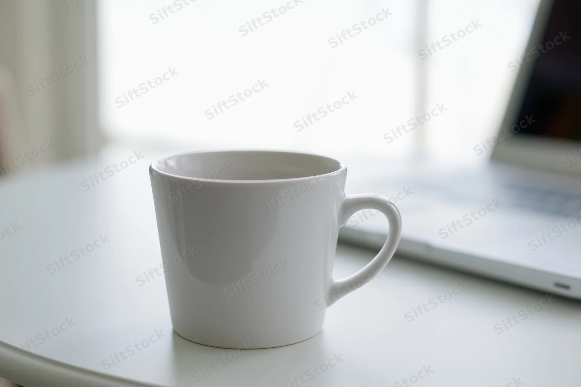 White ceramic coffee mug on a white desk beside a laptop in a bright indoor workspace