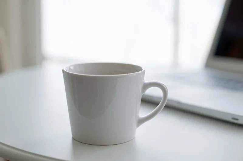 White ceramic coffee mug on a white desk beside a laptop in a bright indoor workspace
