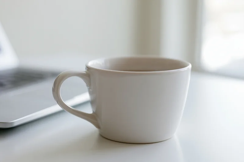 White ceramic coffee mug on a white desk beside a laptop in a bright indoor workspace