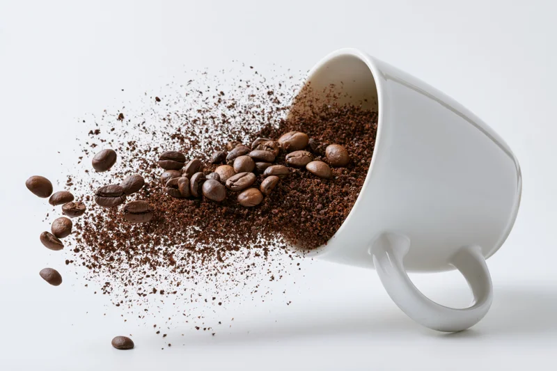 White ceramic coffee mug tipped sideways spilling roasted beans and ground coffee midair against white background