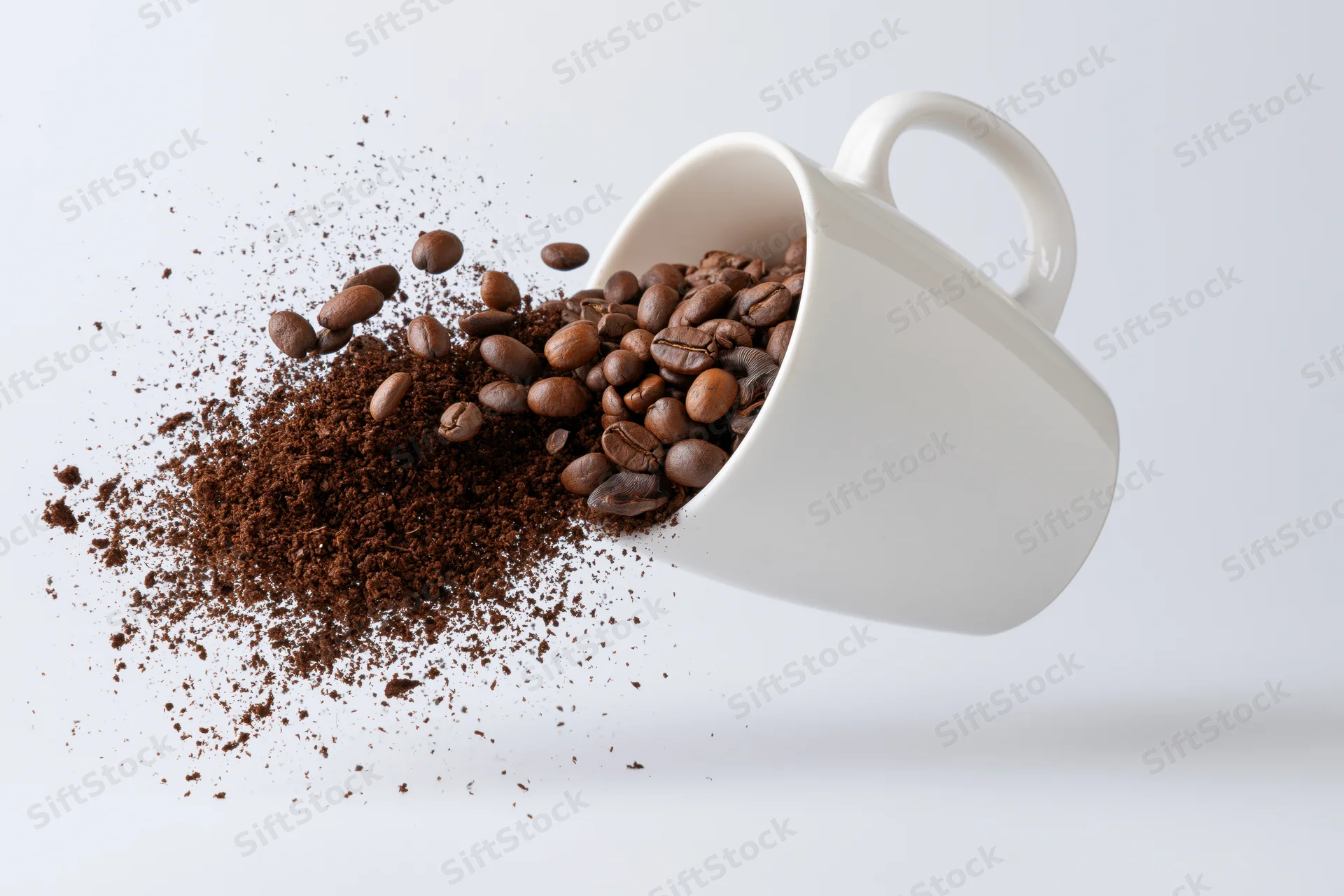 White ceramic coffee mug tipped sideways midair spilling roasted beans and ground coffee against plain white background