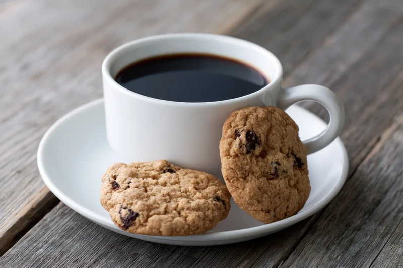 White ceramic cup of black coffee with chocolate chip cookies on a matching saucer on wooden table