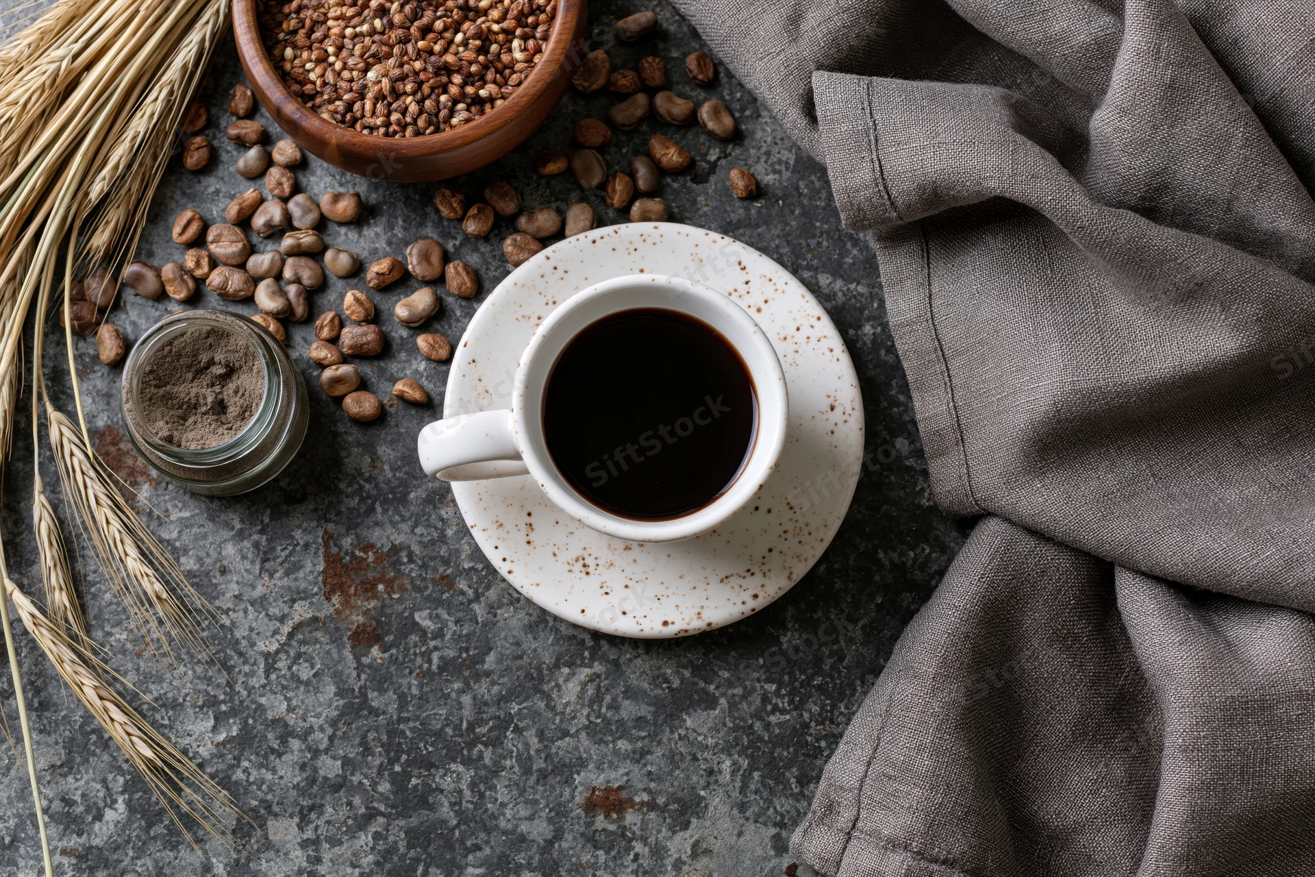 White ceramic cup of black coffee on saucer with coffee beans, wheat stalks, and linen cloth on textured stone surface
