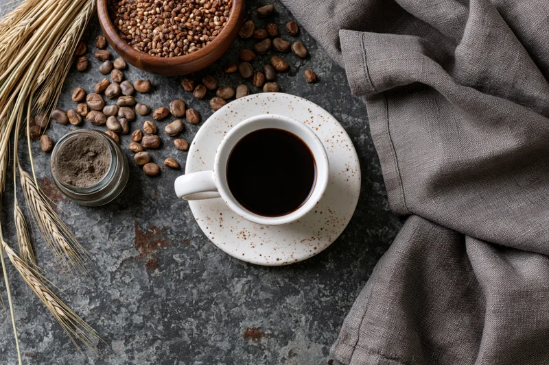 White ceramic cup of black coffee on saucer with coffee beans, wheat stalks, and linen cloth on textured stone surface