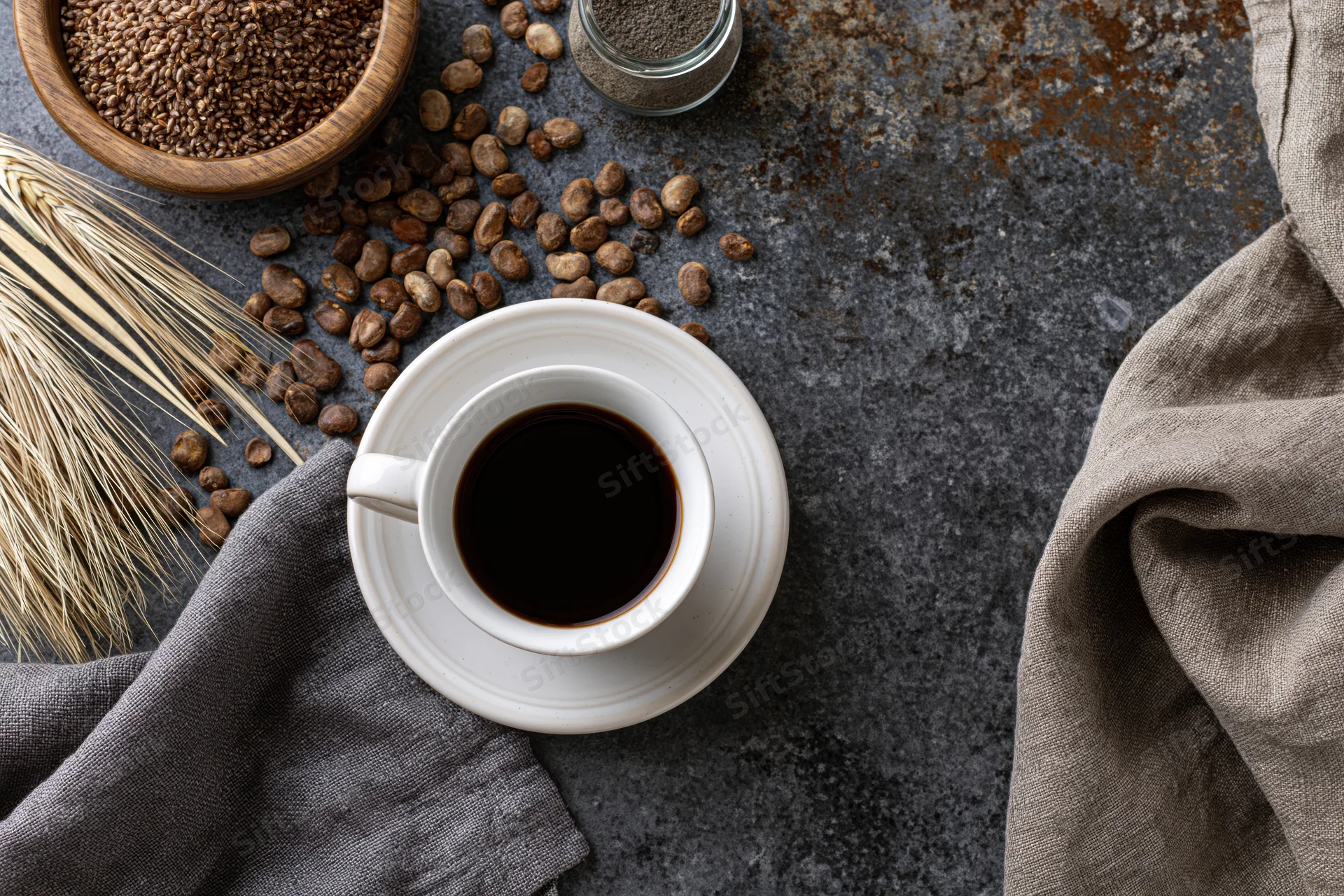 White ceramic cup of black coffee on saucer with roasted beans and linen cloth on dark textured surface