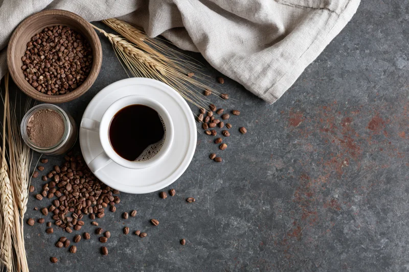 White ceramic cup of black coffee on saucer with scattered beans and wheat stalks on dark stone surface