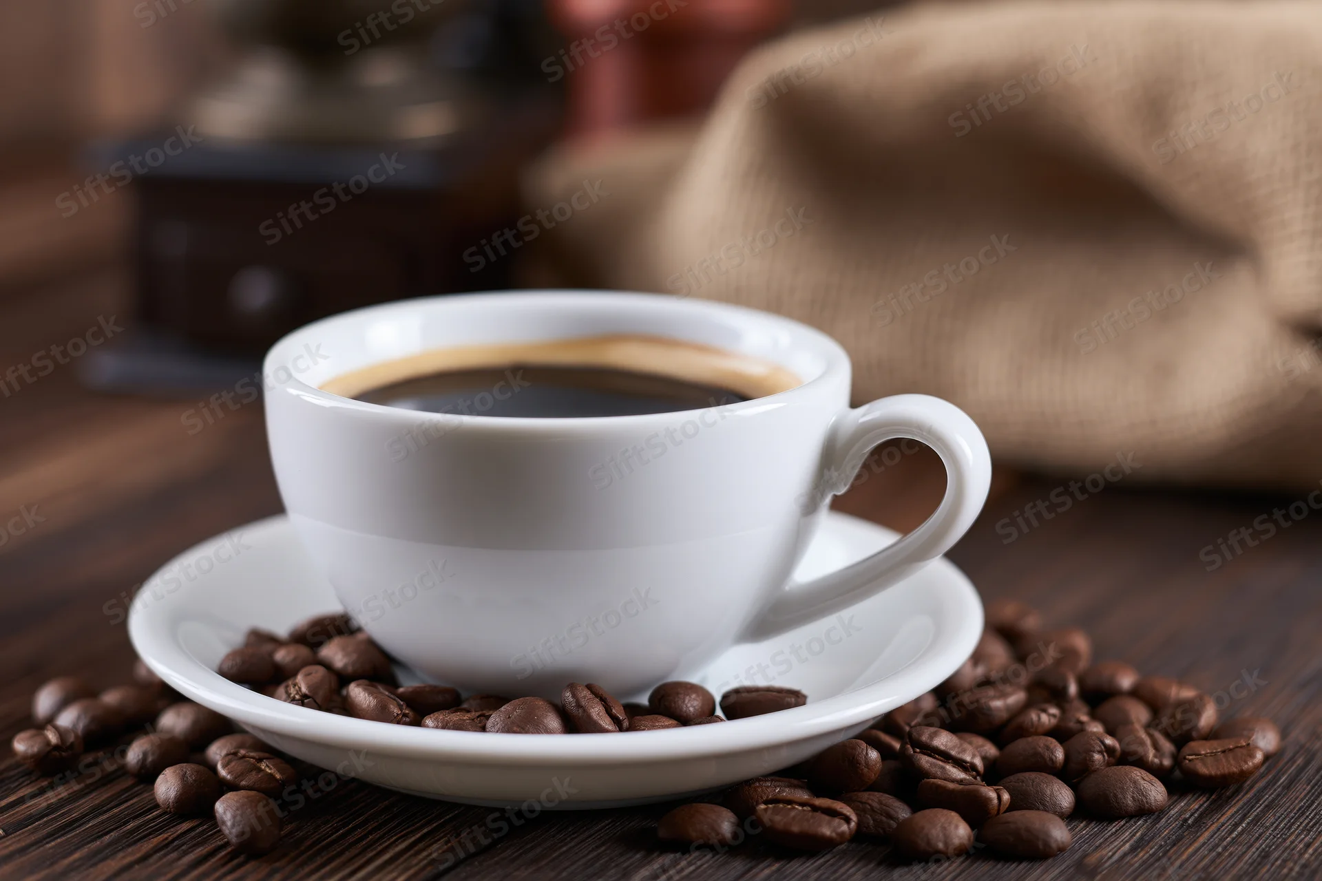 White ceramic coffee cup with foam ring on saucer surrounded by roasted beans on wooden table