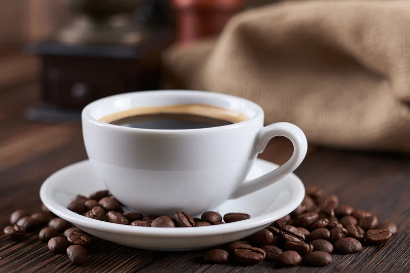 White ceramic coffee cup with foam ring on saucer surrounded by roasted beans on wooden table