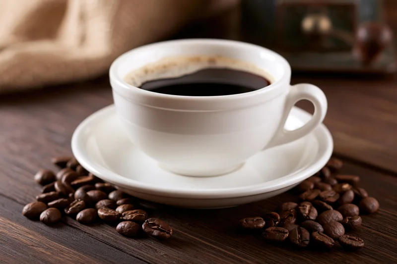 White ceramic cup of black coffee with foam ring on saucer surrounded by roasted beans on dark wooden table