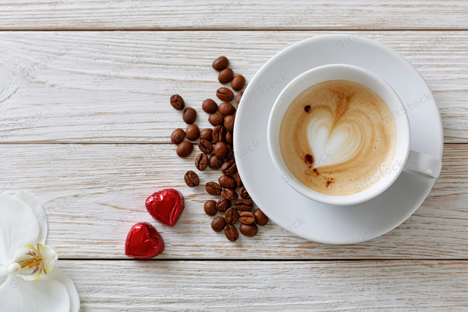 Cappuccino with heart-shaped foam art on a white wooden table beside coffee beans, red heart candies and a white orchid flower