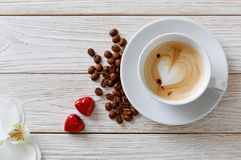 Cappuccino with heart-shaped foam art on a white wooden table beside coffee beans, red heart candies and a white orchid flower