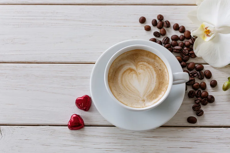 Cappuccino in a white ceramic cup with heart-shaped foam art on a wooden table surrounded by coffee beans, red chocolate hearts, and a white orchid