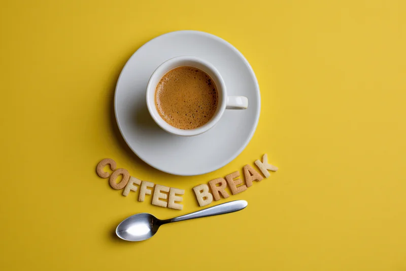 Espresso cup on saucer with cookie letters spelling coffee break and spoon on yellow surface
