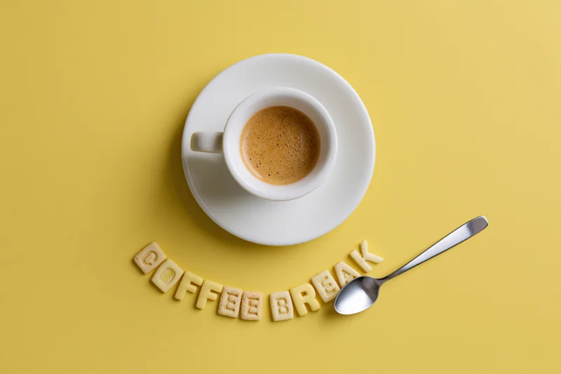 White ceramic espresso cup on saucer with biscuit letters spelling coffee break and spoon on yellow background