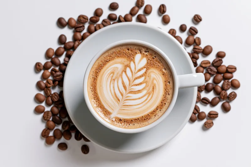 White ceramic cup of latte with leaf foam art surrounded by roasted coffee beans on white surface