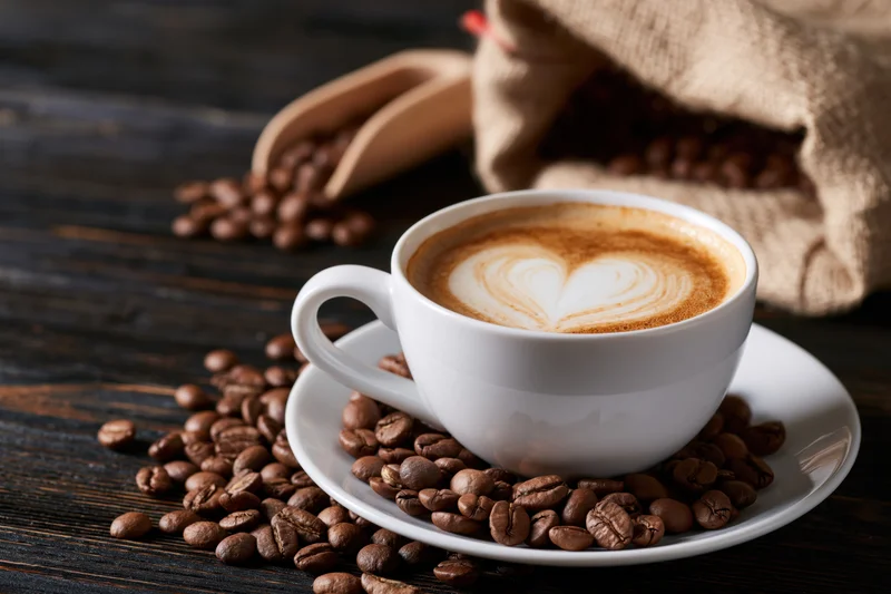 White ceramic cup of latte with heart-shaped foam on saucer surrounded by coffee beans on dark wooden table
