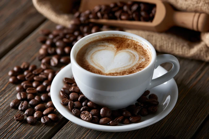 White ceramic cup of latte with heart-shaped foam on saucer surrounded by coffee beans on wooden table